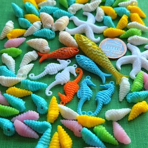 May include: A collection of colorful, miniature ocean-themed charms and beads. The assortment includes various shapes like seashells, starfish, seahorses, and fish, in shades of yellow, pink, blue, green, and white. A US penny is included for size reference.