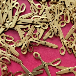May include: A large pile of gold-colored miniature scissors on a pink background. The scissors are all the same size and shape, and they are arranged in a random pattern.