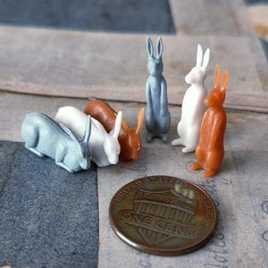 May include: A collection of small, colorful rabbit figurines in various poses. The rabbits are in shades of gray, white, and orange. A copper one-cent coin is included for scale. The figurines are likely for decorative purposes.