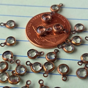 May include: Close-up of small, clear faceted glass charms with a copper-colored metal setting and loop. The charms are scattered on a lined paper surface, with a penny for size comparison. The charms have a vintage aesthetic.