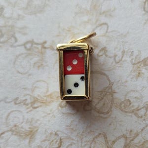 May include: A gold-toned charm featuring a domino design. The domino is divided into two sections: the top half is red with three white dots, and the bottom half is white with three black dots. The charm has a small loop for attachment.