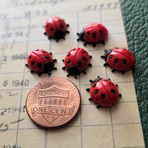 May include: Six small, red ladybug-shaped buttons with black spots and black heads, arranged alongside a copper United States one-cent coin for scale. Each ladybug features a rounded, glossy red shell with distinct black markings.