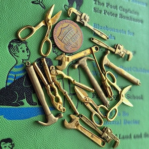 May include: A collection of miniature gold-colored tools, including scissors, wrenches, and hammers, arranged on a green background. A US one-cent coin is also present. The tools are likely charms or decorative items.