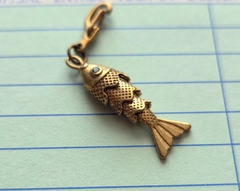 May include: A gold-coloured fish charm with a textured body and a small blue accent on the eye. The charm is attached to a clasp. The background is a light blue paper with blue lines.