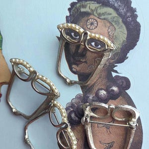 May include: Three vintage eyeglasses-shaped brooches. Two are silver-toned with faux pearl accents, and one is gold-toned with pearl accents. The brooches are arranged on a vintage illustration background.