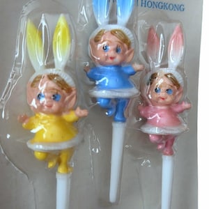 May include: Three plastic bunny-eared figurines in yellow, blue, and pink dresses. Each figure has a white collar and a white stem. The packaging reads "STY... ! HONGKONG".