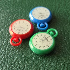 May include: Three small round plastic pocket watch charms in red, blue, and green. Each charm features a white clock face with black numerals and the text Elgin printed in the center. Each piece includes a small circular loop at the top.