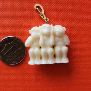 May include: A white ceramic charm of three monkeys, one covering its eyes, one covering its ears, and one covering its mouth. The charm is attached to a gold-colored metal loop.