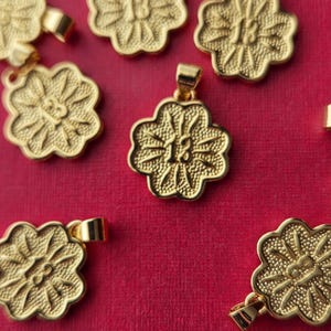 May include: Gold-colored pendants with a floral design and the number 13 are scattered on a red background. Each pendant has a loop for attaching to a chain or bracelet. The pendants are detailed with textured surfaces.