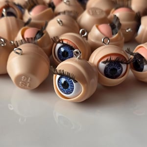 May include: A collection of doll eyes with blue irises, long black eyelashes, and a light peach-colored eyelid. Each eye has a small metal loop attached. The eyes are set against a neutral background.