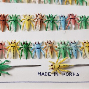 May include: Rows of colorful insect pins in various colors, including red, green, yellow, and blue. Each pin features a detailed insect design with red eyes and a pin backing. The text "MADE IN KOREA" is visible.