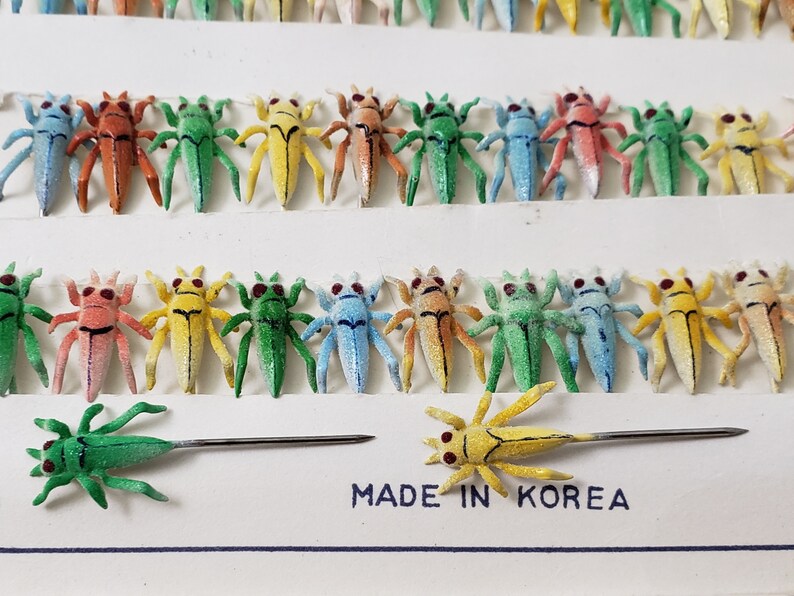 May include: Rows of colorful insect pins in various colors, including red, green, yellow, and blue. Each pin features a detailed insect design with red eyes and a pin backing. The text "MADE IN KOREA" is visible.
