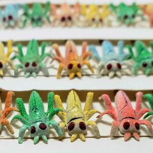 May include: A collection of colorful, glittery insect figurines in various shades of blue, green, yellow, orange, and pink. Each figurine has multiple legs and red eyes, arranged in neat rows. The background is a plain white surface.