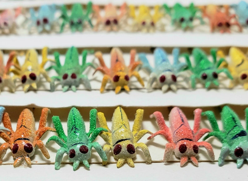 May include: A collection of colorful, glittery insect figurines in various shades of blue, green, yellow, orange, and pink. Each figurine has multiple legs and red eyes, arranged in neat rows. The background is a plain white surface.