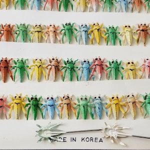 May include: Rows of colorful, miniature insect figurines in various colors, including green, yellow, orange, and blue. Each insect has a detailed design with tiny legs and antennae. The text "MADE IN KOREA" is visible at the bottom.