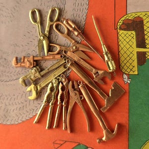 May include: A collection of miniature gold-toned metal tool charms including scissors, an adjustable wrench, a pipe wrench, pliers, a hammer, a screwdriver, and a nutcracker. Each small tool features a metallic finish and intricate detailing.