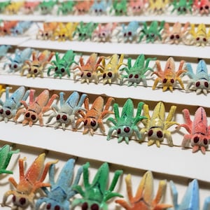May include: Rows of colorful, miniature insect figurines in various colors including green, orange, yellow, and blue. Each insect has six legs, a segmented body, and red eyes. The figurines are arranged on white cardstock.