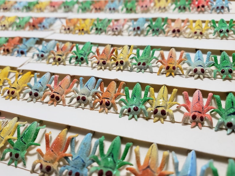 May include: Rows of colorful, miniature insect figurines in various colors including green, orange, yellow, and blue. Each insect has six legs, a segmented body, and red eyes. The figurines are arranged on white cardstock.
