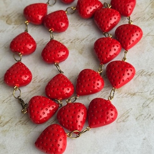 May include: A necklace featuring bright red strawberry-shaped beads. Each bead has a textured surface with small, evenly spaced dots. The beads are linked together with gold-colored metal chain.