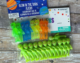 Peut inclure: Plusieurs figurines d'aliens phosphorescents de couleurs variées, vert, bleu, jaune et orange. L'emballage affiche "Glow in the Dark Aliens" et "Party Favors". Une mise en garde concernant les petites pièces est également présente.