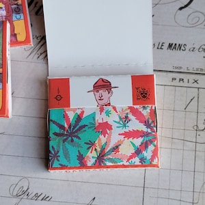 May include: A vintage pack of cigarette papers with a red, white, and green design. The front of the pack features a cartoon image of a man wearing a hat and a red shirt. The text "CIGARETTE PAPER" is printed on the side of the pack.
