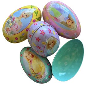 May include: A collection of five Barbie-themed egg-shaped tins. The tins feature images of Barbie, floral patterns, and the Barbie logo. Colors include yellow, pink, purple, and teal. One tin is open, revealing the interior.