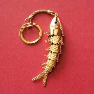 May include: A gold-colored fish-shaped keychain with a blue gem eye. The fish has detailed scale and fin designs. The keychain includes a ring and a decorative, flexible connector. The background is a solid red.