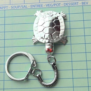 May include: A silver-colored turtle keychain with red eyes. The turtle has a detailed shell design and is attached to a metal chain and keyring. The background is a green and white grid with the text "Guest Check".