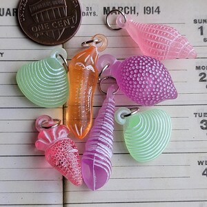 May include: Seven colorful plastic charms with metal rings. The charms are shaped like a seashell, a strawberry, a corn cob, a clam, a seashell, a lemon, and a seashell. The charms are on a white lined paper with the date March 1914.