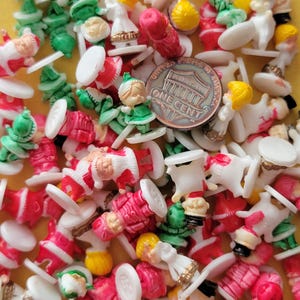 May include: A large collection of small plastic figurines of Christmas characters, including Santa Claus, elves, and snowmen. The figurines are all in various colors, including red, green, white, and yellow. The figurines are all standing on small white bases. There is a US penny in the image for scale.