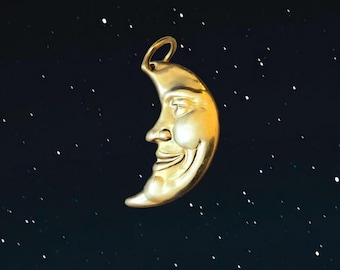 May include: A gold-toned pendant shaped like a crescent moon with a human-like face featuring a closed eye, a prominent nose, and a smiling mouth. The pendant includes a circular bail at the top for attachment to a chain or cord.