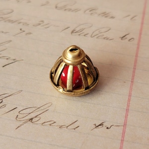 May include: A gold-toned metal cage with a red die inside. The cage is shaped like a bell and has a decorative design.