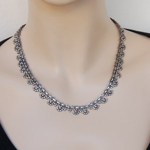 Può includere: Una collana d'argento con un designo delicato e intricato. La collana è realizzata con piccole perline rotonde disposte in un motivo di gocce a cascata.