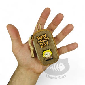 May include: A small, rectangular, tan-colored zippered pouch featuring a beige paracord zipper pull. The front displays a yellow and black rubberized patch with the text "TODAY IS A GOOD DAY" above a cartoon character with spiky yellow hair.