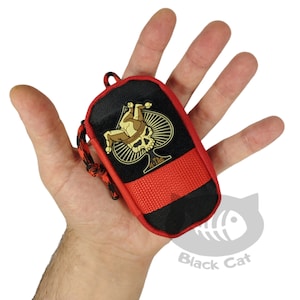 May include: A small rectangular black and red zippered pouch featuring a gold-colored patch of a skull wearing a jester hat inside a spade shape. The pouch has a red paracord zipper pull and a red nylon strap across the lower front section.