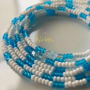 May include: Strands of waist beads in a repeating pattern of white and turquoise blue beads. The beads are small and round, with a glossy finish. The beads are strung together to form a flexible strand. The background is a soft, blurred white.