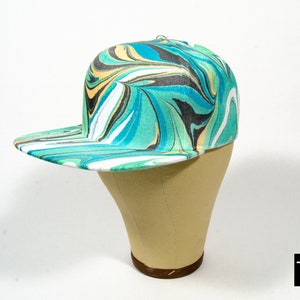 May include: A green, blue, white and black marbled baseball cap with a flat brim. The cap is on a tan mannequin head. The text "12-L" is in the bottom right corner of the image.