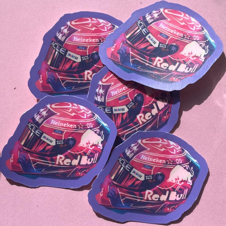 May include: A collection of five stickers featuring a racing helmet design. The helmets are primarily red with white and blue accents, and the Red Bull logo is prominently displayed. The stickers have a purple border.
