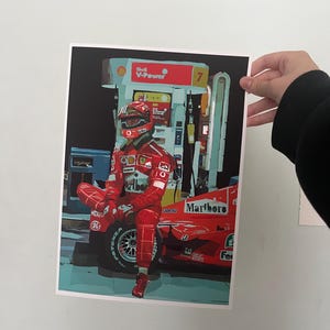May include: A red Formula One race car with the Marlboro logo on the side. A race car driver wearing a red racing suit and helmet is sitting on the car. The car is parked at a gas station with a Shell V-Power sign.