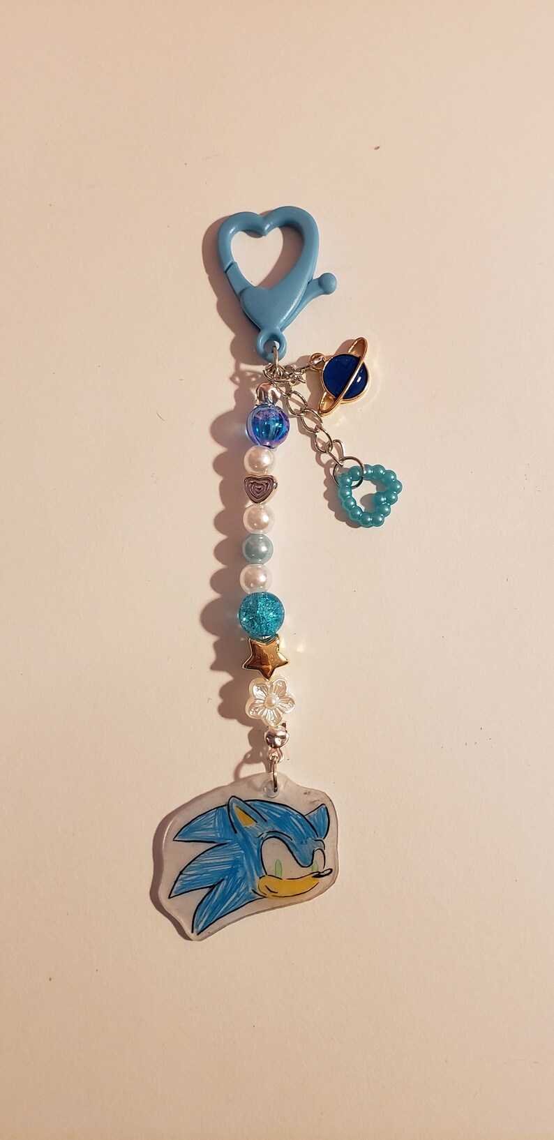 May include: A blue and white beaded keychain with a blue and gold planet charm, a blue heart charm, and a blue and white Sonic the Hedgehog charm.