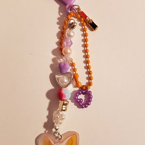 May include: A purple heart-shaped keychain with a charm of a cartoon character with yellow ears and blue eyes. The keychain has a variety of beads, including purple, white, and pink hearts, clear beads, and a small orange chain.