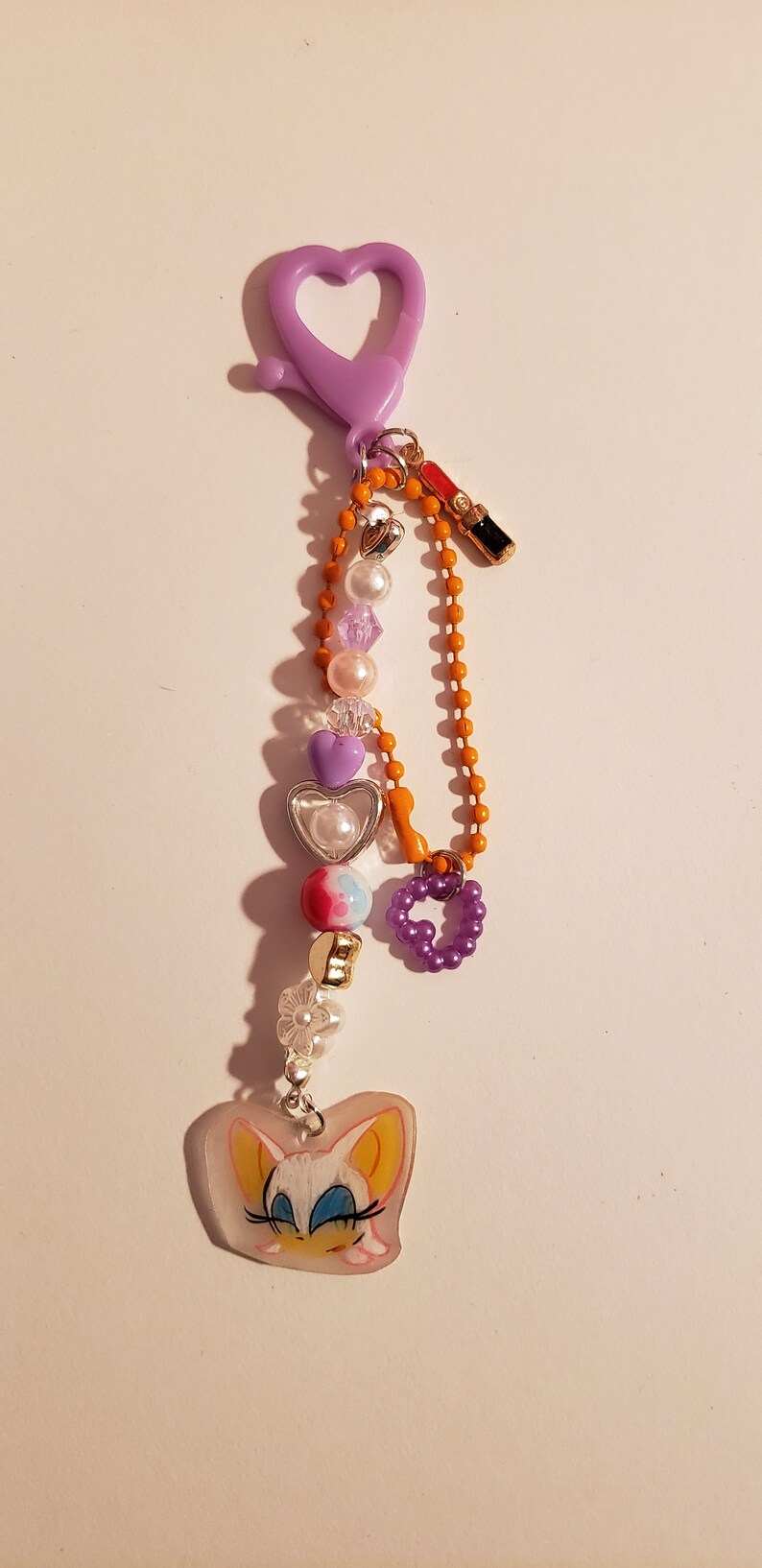 May include: A purple heart-shaped keychain with a charm of a cartoon character with yellow ears and blue eyes. The keychain has a variety of beads, including purple, white, and pink hearts, clear beads, and a small orange chain.