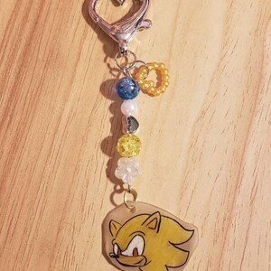 May include: A yellow and white keychain featuring a cartoon character with a red nose and blue eyes. The keychain has a silver heart-shaped clasp and a string of beads in blue, white, and yellow.