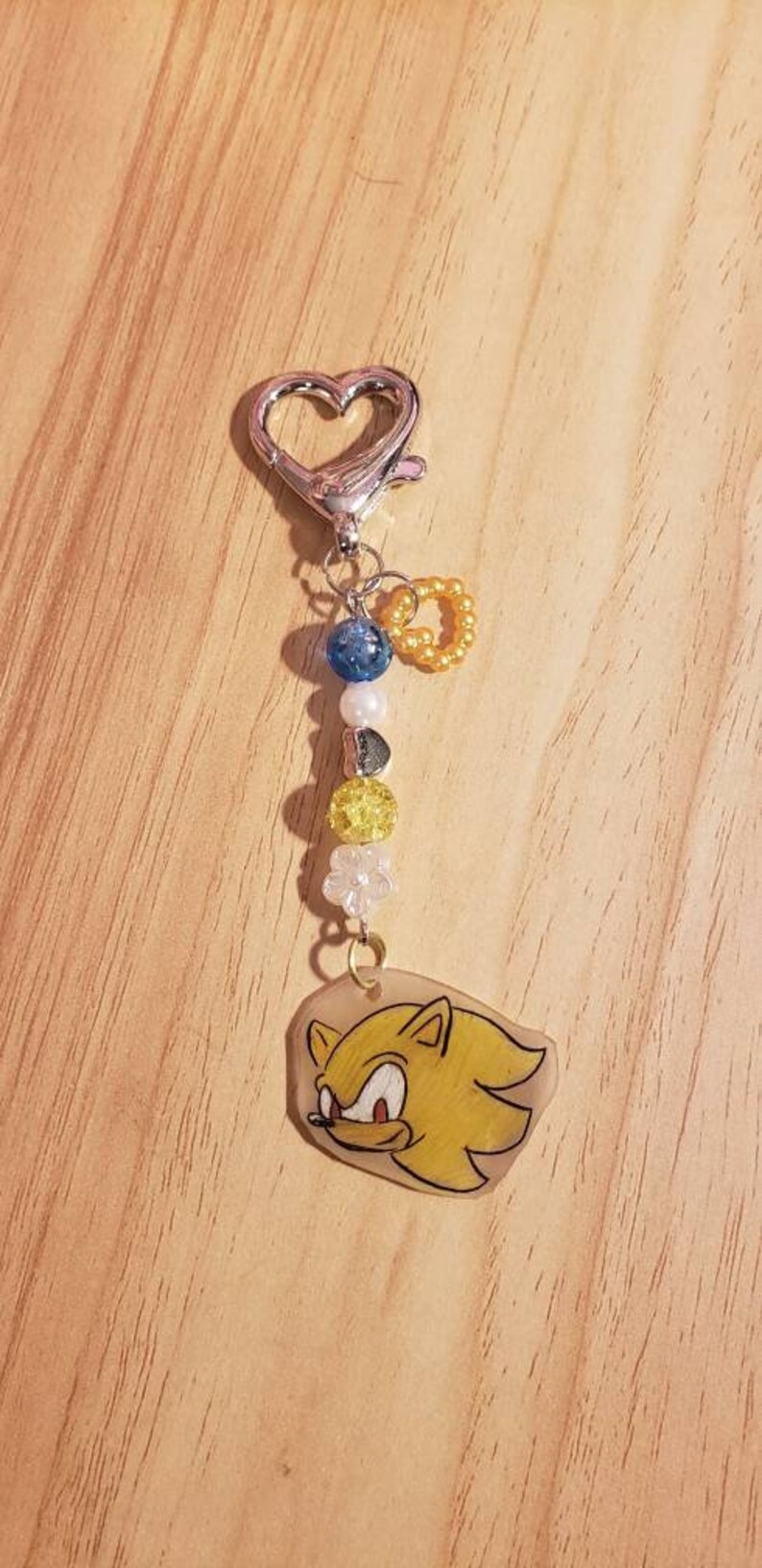 May include: A yellow and white keychain featuring a cartoon character with a red nose and blue eyes. The keychain has a silver heart-shaped clasp and a string of beads in blue, white, and yellow.