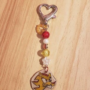 May include: A silver heart-shaped keychain with a red, white, and yellow beaded charm featuring a cartoon character with a yellow and red design.