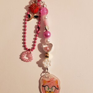 May include: Pink and white beaded keychain with a heart-shaped charm, a rose charm, and a clear charm with a drawing of Amy Rose from Sonic the Hedgehog.