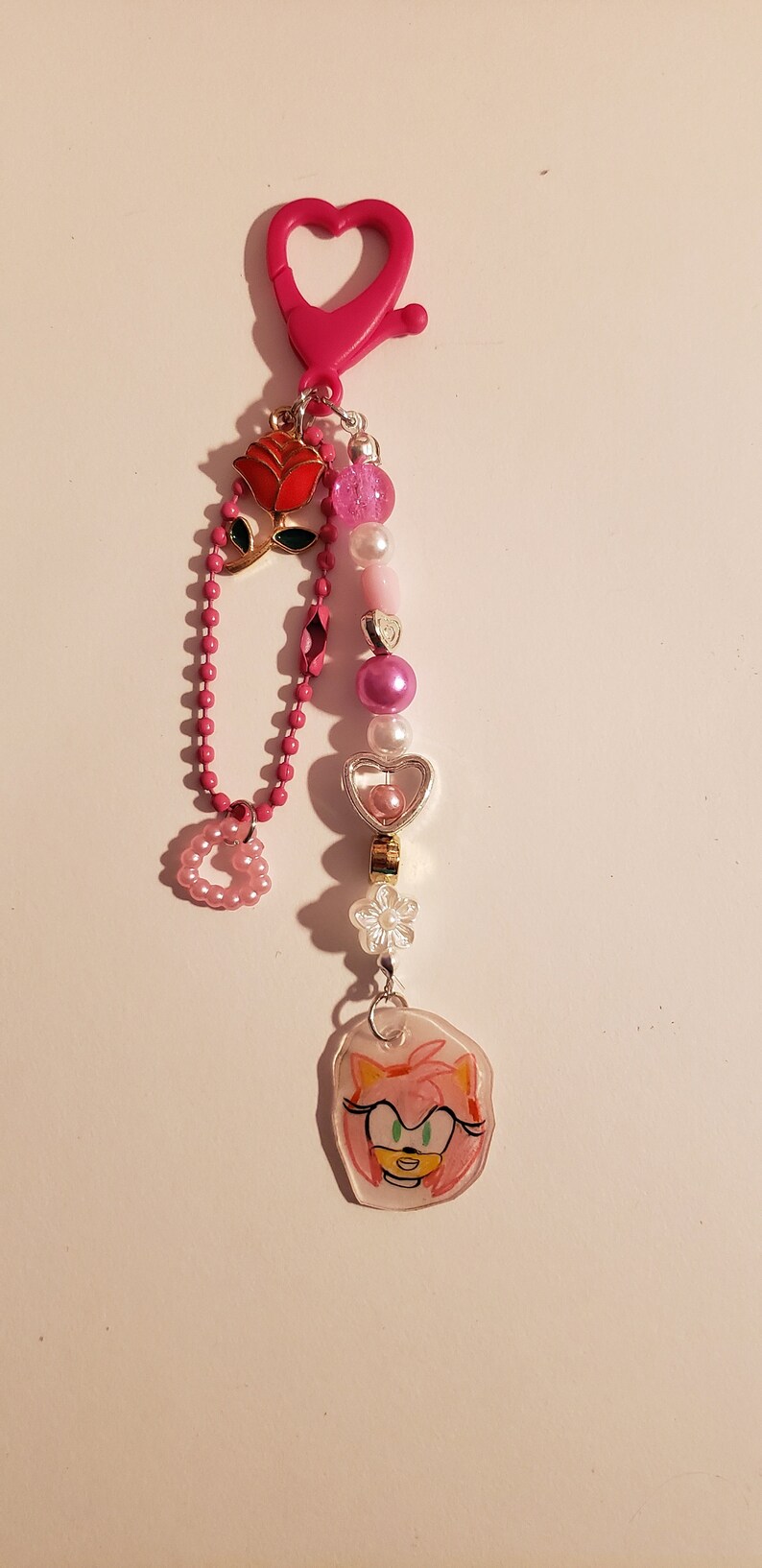 May include: Pink and white beaded keychain with a heart-shaped charm, a rose charm, and a clear charm with a drawing of Amy Rose from Sonic the Hedgehog.