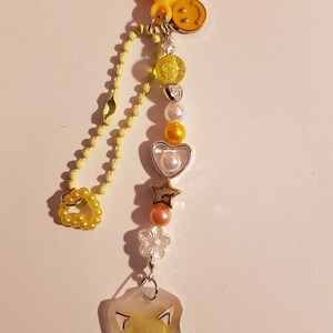 May include: A yellow heart-shaped keychain with a yellow chain and various charms, including a smiley face, hearts, stars, and a clear charm with a cartoon image of a yellow fox.