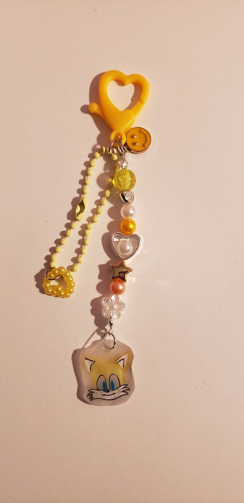 May include: A yellow heart-shaped keychain with a yellow chain and various charms, including a smiley face, hearts, stars, and a clear charm with a cartoon image of a yellow fox.