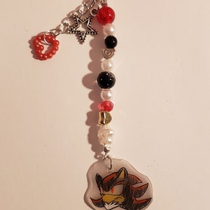 May include: A black keychain with a red heart, a silver star, and a clear charm with a drawing of Shadow the Hedgehog. The keychain is decorated with red, white, and black beads.